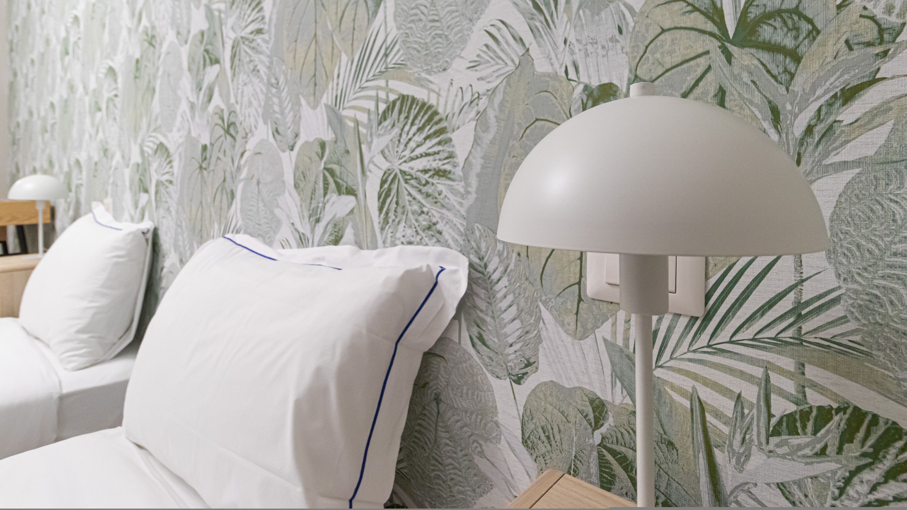 A bedroom with a bed, white pillows, a nightstand, a lamp, and a wall with a tropical leaf pattern.