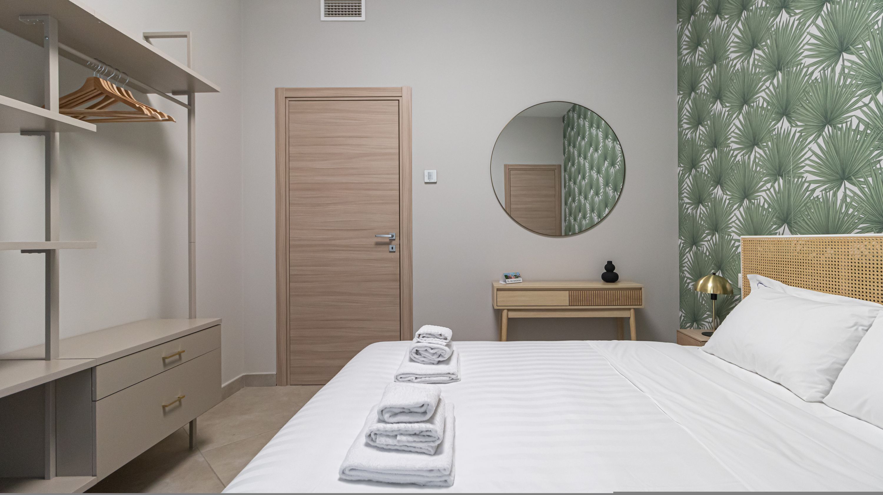 Ein Hotelzimmer mit Bett, Holzschrank, rundem Spiegel, Nachttisch und einer Wand mit Blätermotiv.