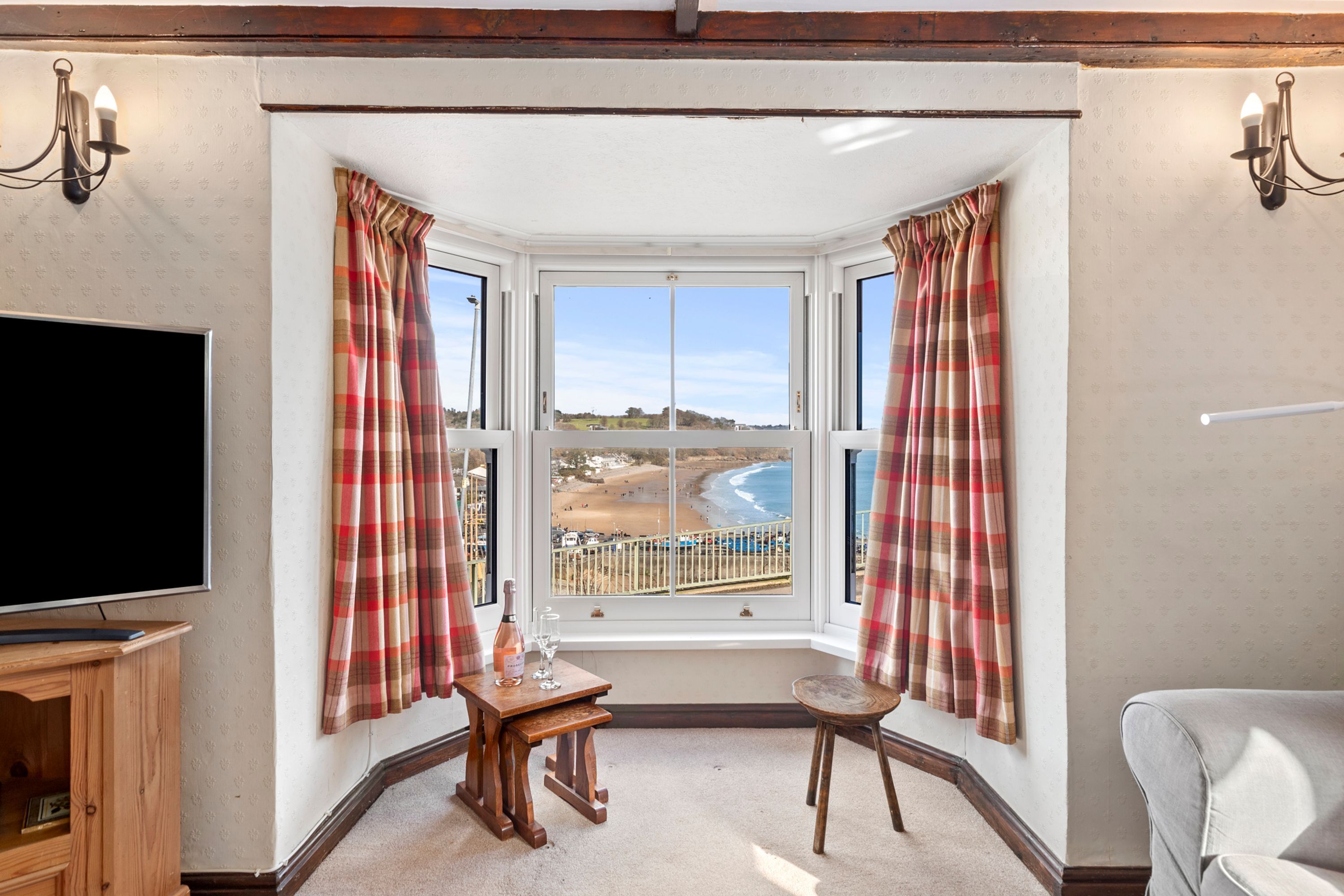 A corner room with a bay window overlooking a beach, a television, a small wooden table with a bottle and glasses, and a chair.