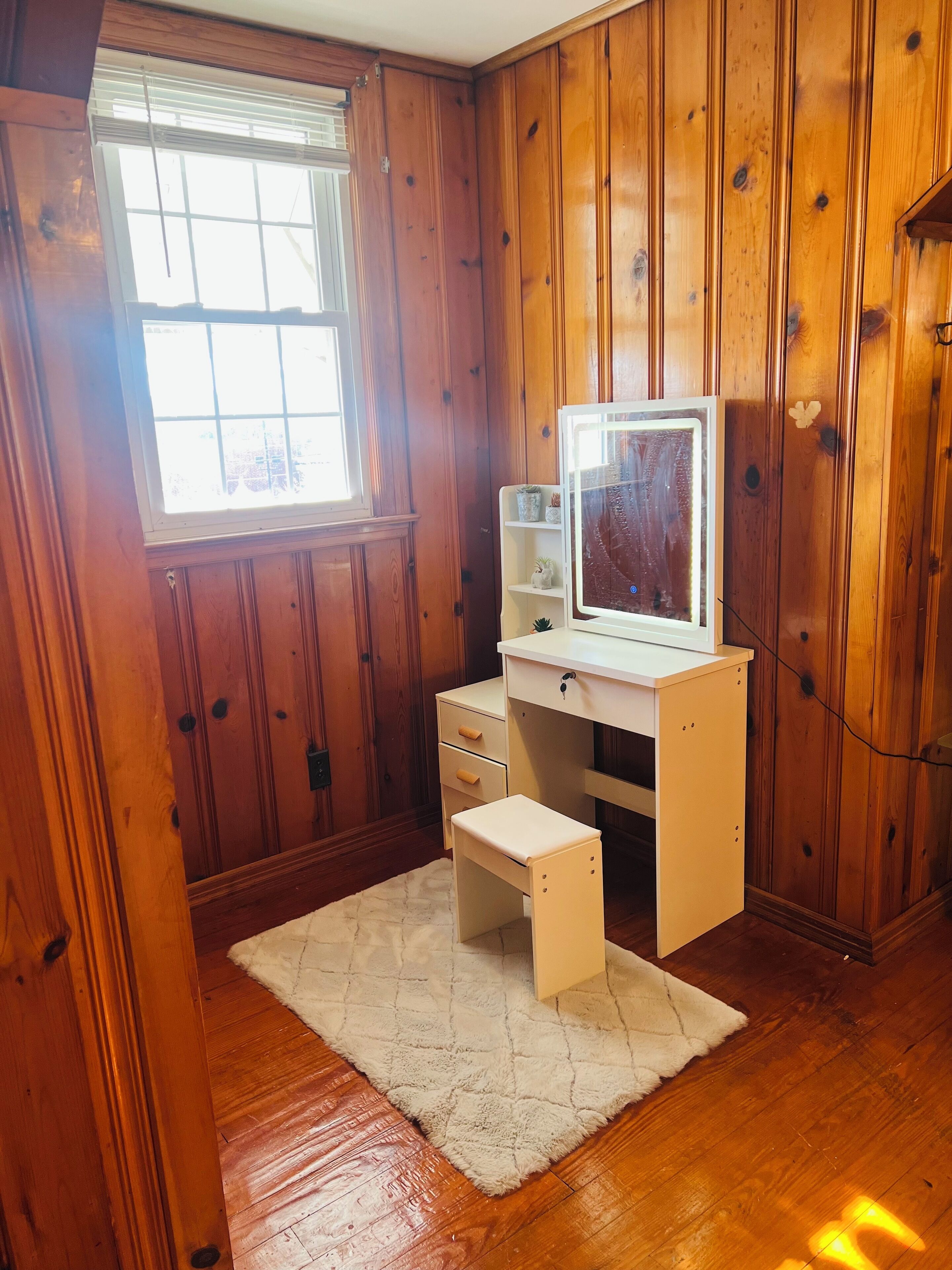 Une petite pièce aux murs lambrissés, comprenant un bureau, une chaise, une fenêtre et un tapis blanc.