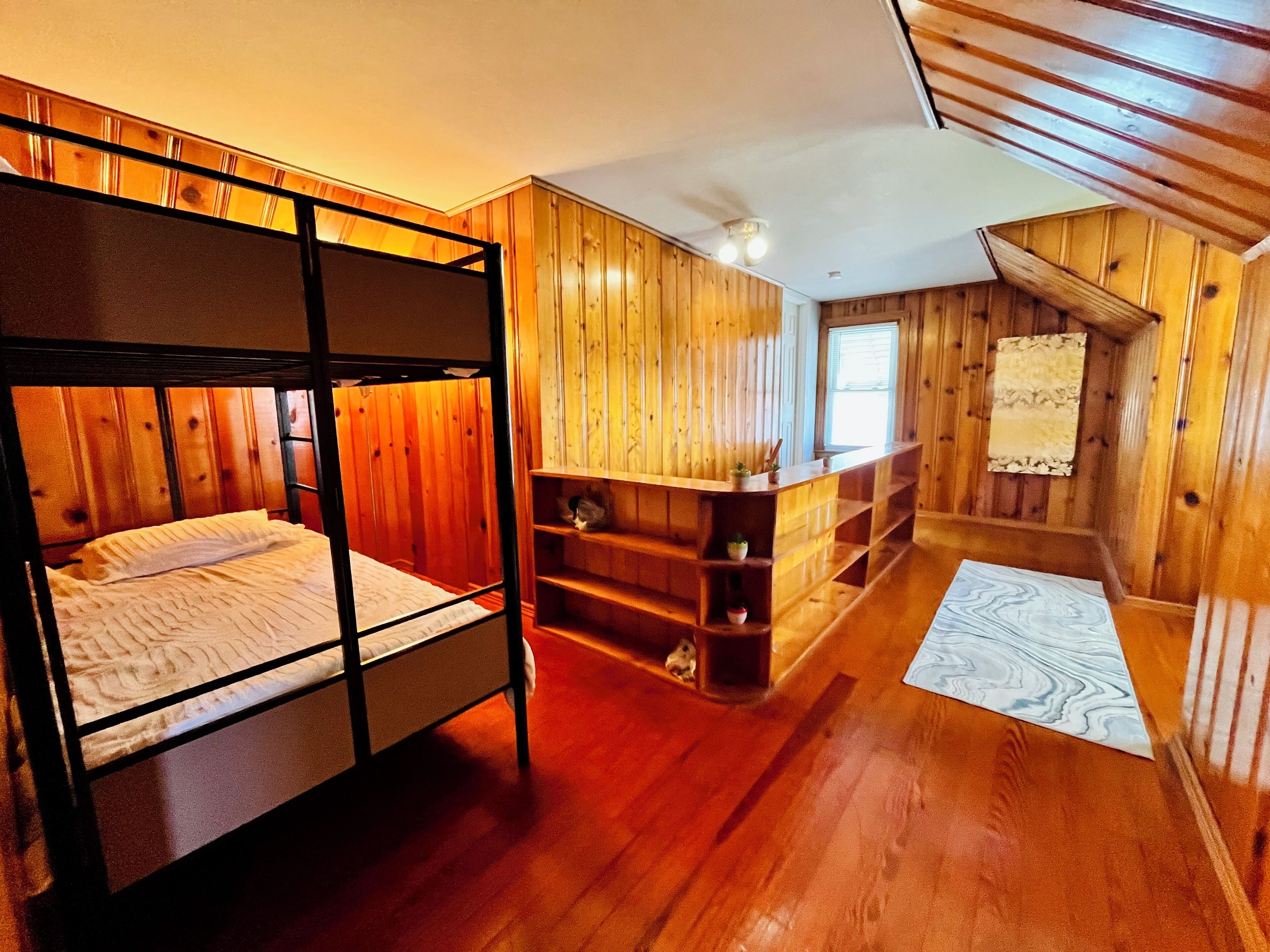 Une chambre avec des lits superposés, des murs en bois et un plafond en pente. On y trouve une petite étagère avec des plantes et une fenêtre avec des stores.