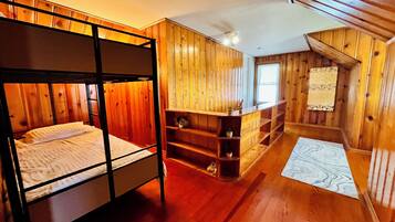Une chambre avec des lits superposés, des murs en bois et un plafond en pente. On y trouve une petite étagère avec des plantes et une fenêtre avec des stores.