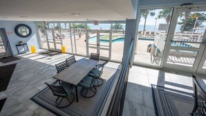 Un espacio interior amplio con una gran mesa y sillas, ventanas grandes que ofrecen vistas a una piscina y una playa, y un cielo despejado.