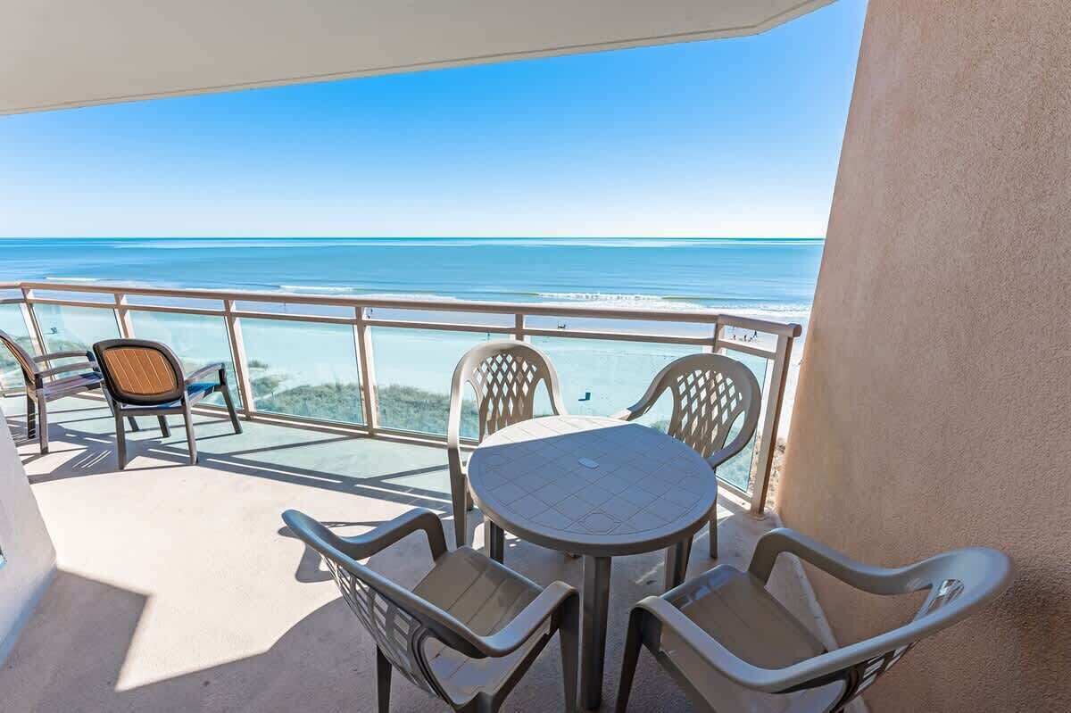 A balcony with a table and chairs, overlooking a beach with clear blue water.
