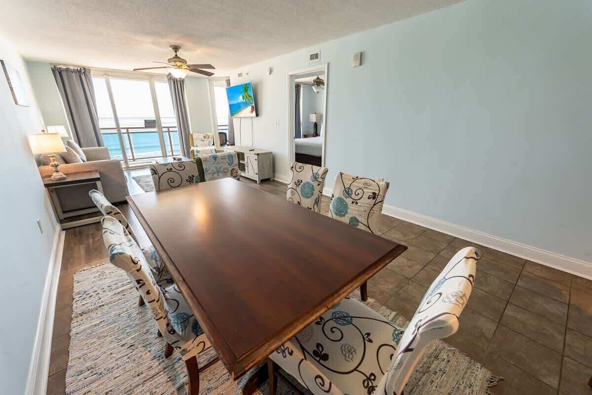 A dining area with a large wooden table, patterned chairs, and a view of the sea through large windows.