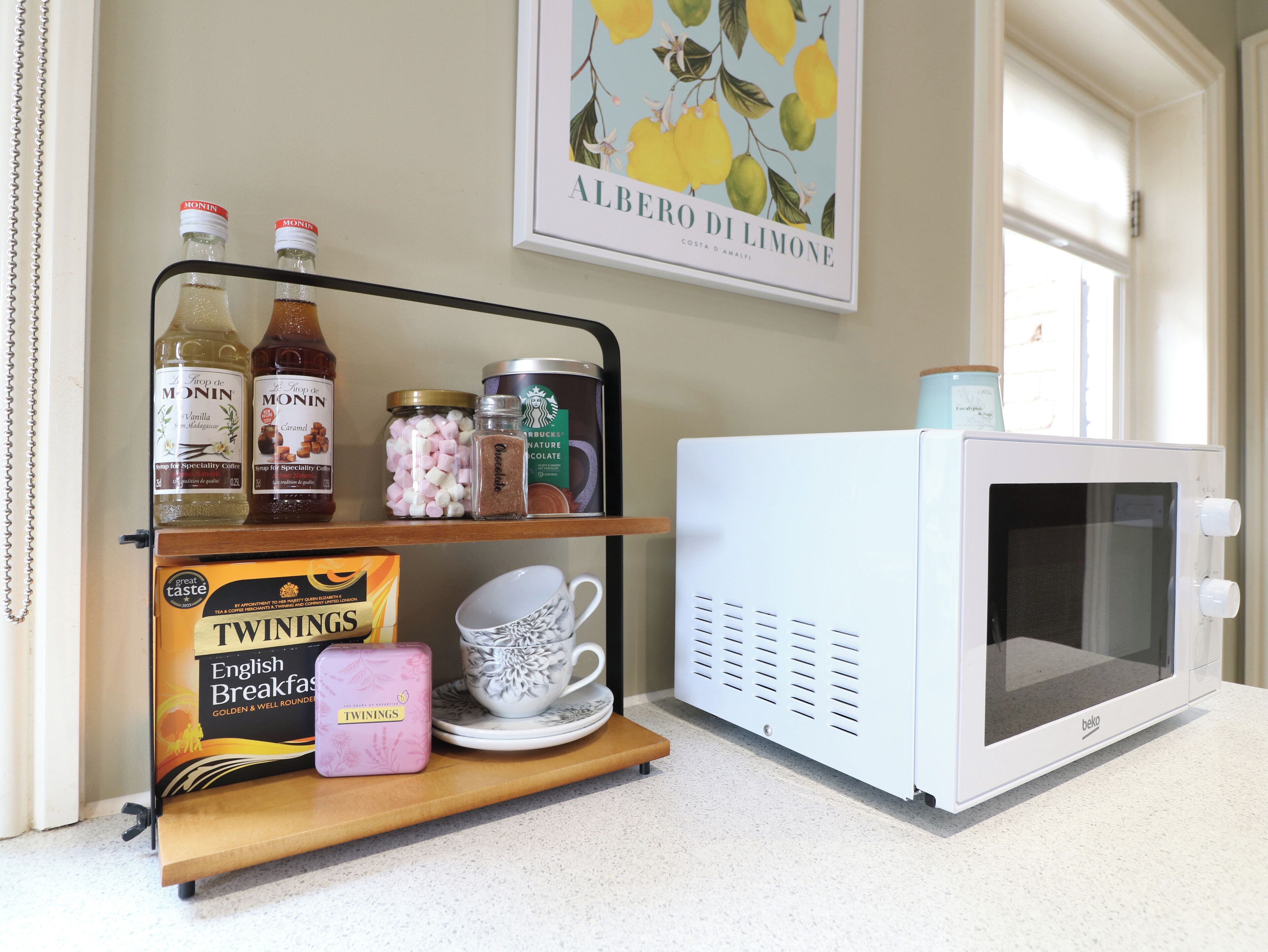 En köksbänk med en mikrovågsugn, en hylla med diverse föremål som flaskor och en ask med Twinings English Breakfast-te, och en inramad tavla på väggen.