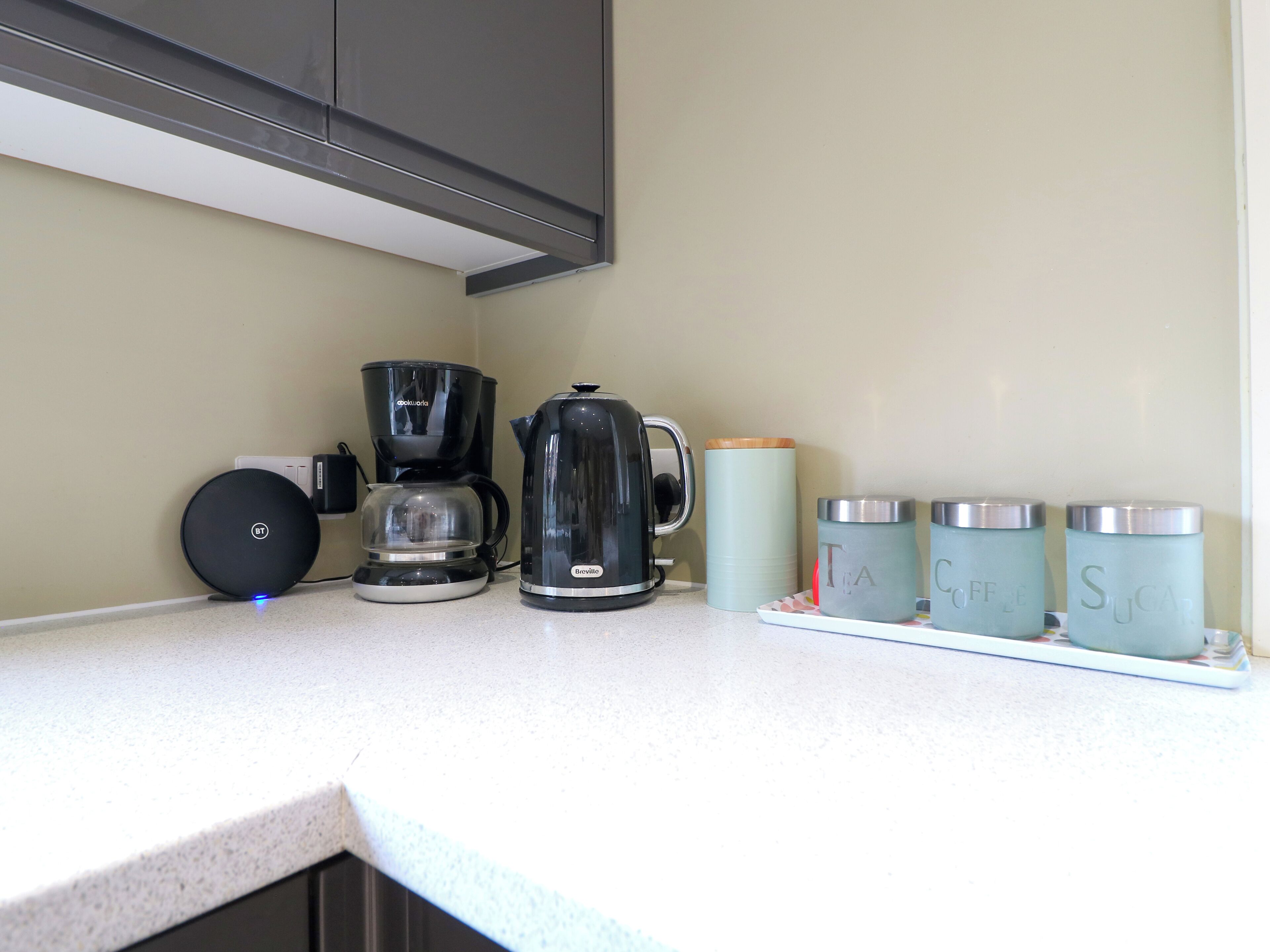 A kitchen counter with a coffee maker, a kettle, and three containers labeled with tea and sugar.