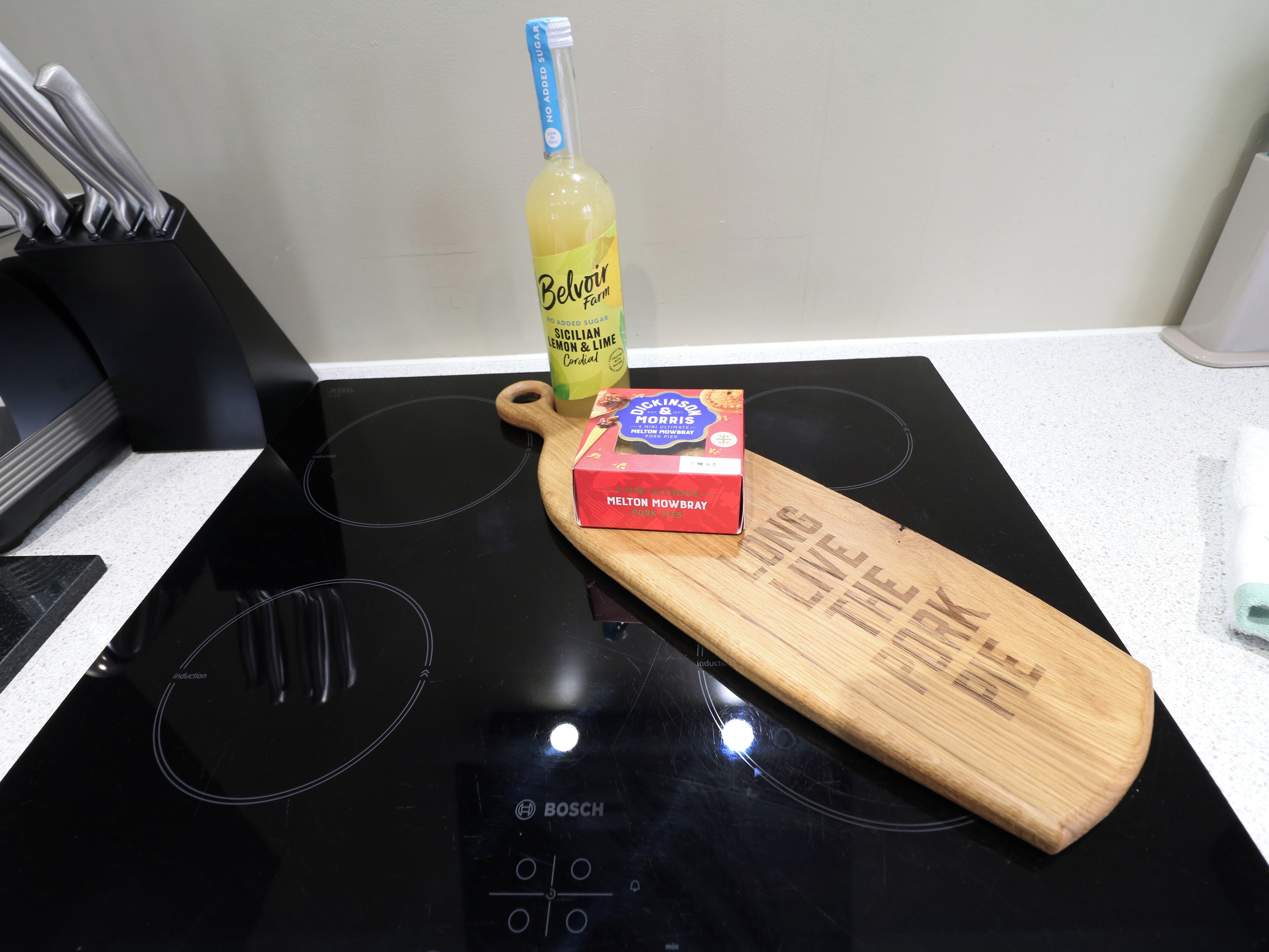 A kitchen with a black induction cooktop, a wooden cutting board, a bottle of Belvoir Farm lemon and lime cordial, and a box of Dickinson & Morris butter.