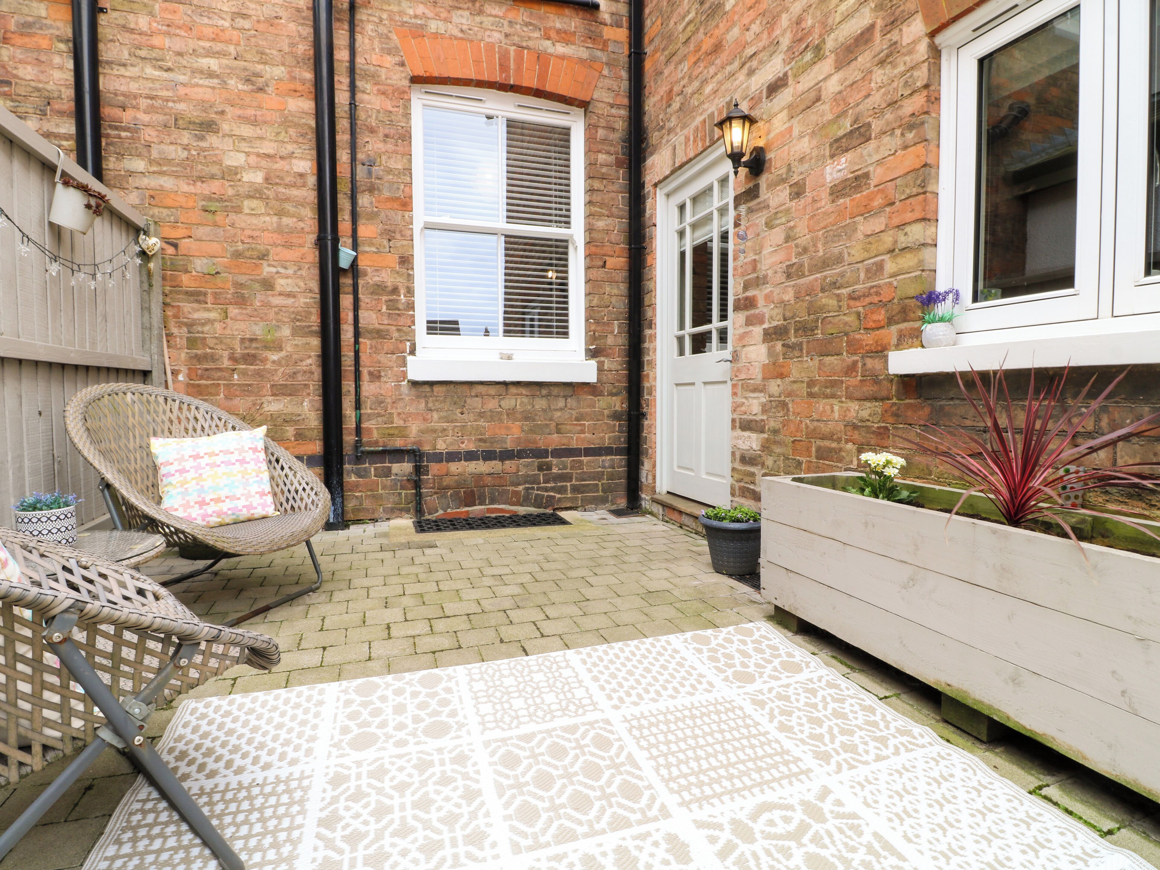 A brick house with a white door, a window with shutters, a wicker chair, a patterned rug, and a planter with plants.