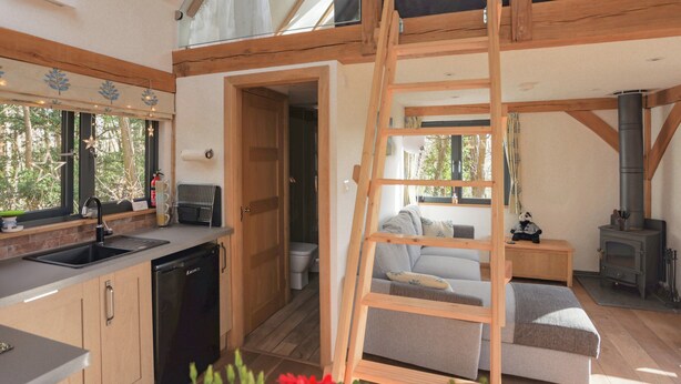 Ein gemütliches Blockhaus mit Schlafzimmer auf dem Dachboden, einer Küche inklusive Spüle und Geschirrspüler, einem Wohnbereich mit Sofa und einer Klapptreppe sowie einem Holzofen.