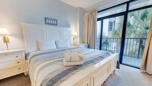 A hotel room with a large bed, a nightstand, a bedside lamp, a window with curtains, and a view of palm trees. - Contemporary 2BR Beach Colony Resort, Pools, View (Myrtle Beach)