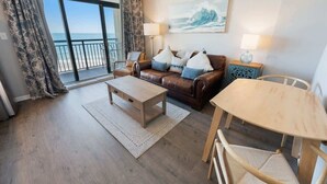 Un salon comprenant un canapé, une table basse et une table à manger, un tableau accroché au mur et une vue sur l’océan par de grandes fenêtres.