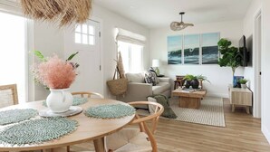 En moderne stue med spiseplads, med et rundt træbord, vævede underlag, en hængende lampe og et stort vindue med gardiner.