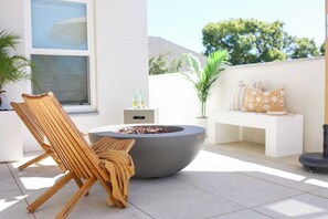 Un espace extérieur moderne, aménagé avec un foyer, des fauteuils de jardin en bois et un banc blanc agrémenté de coussins à motifs.