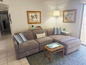 Ein Wohnzimmer mit einem grauen Ecksofa, einem kleinen Holztisch, einem blau gemusterten Teppich und zwei gerahmten Pflanzenbildern an der Wand.