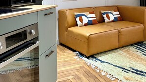 A compact kitchen with a microwave, oven, and a wooden countertop. A brown leather sofa with patterned cushions is placed against a wall, and a herringbone-patterned wooden floor is visible. - Id-home Le Saint Nicolas (Nantes)