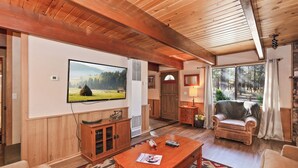 Ein gemütliches Wohnzimmer mit einem großen Flachbildfernseher, Holzmöbeln und Blick ins Freie durch große Fenster.