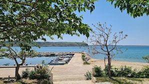 A marina with boats docked, a sandy beach, and a clear view of the sea and distant hills.