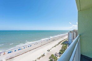Vista da praia a partir de uma varanda, com céu claro, areal e um edifício próximo à costa.