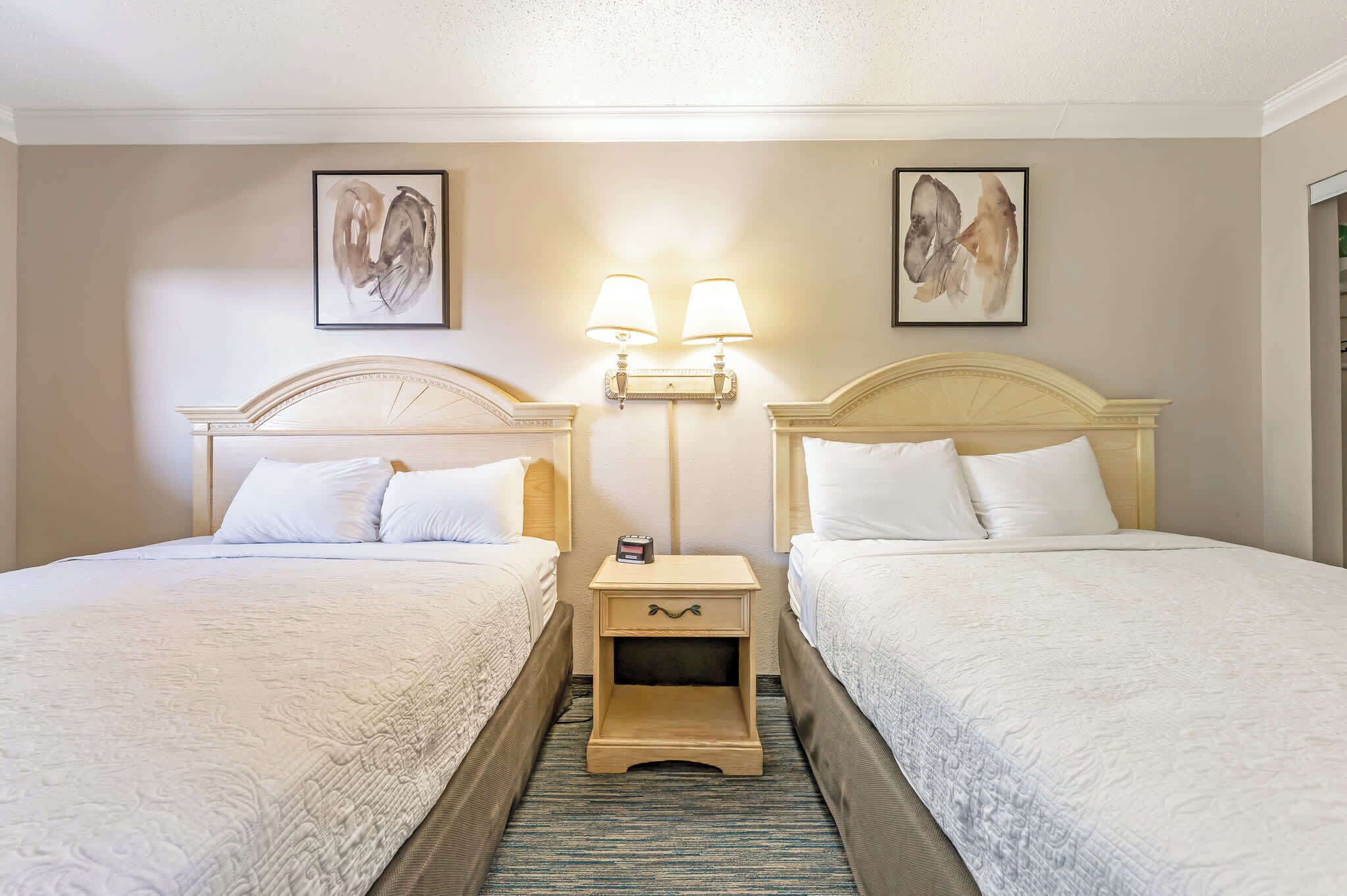 Quarto de hotel com duas camas, uma mesa de cabeceira com abajur e obras de arte nas paredes.