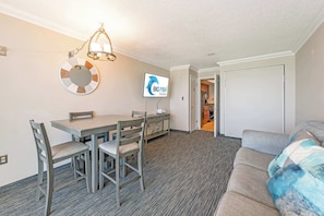 Quarto de hotel com uma mesa de jantar, cadeiras, televisão, sofá e uma porta que dá para outro espaço.