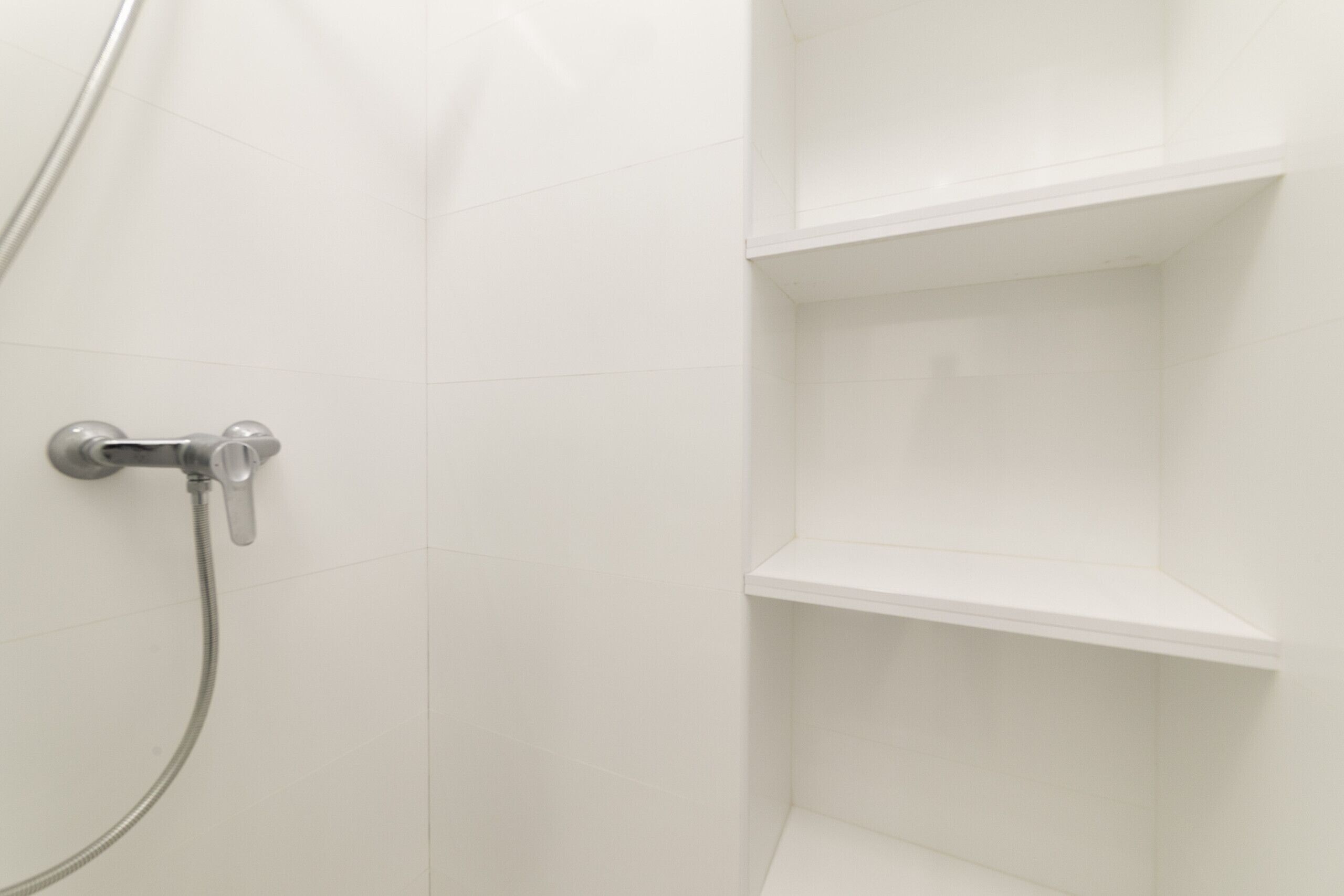 Rincón de una habitación con paredes de azulejos blancos, zona de ducha con grifo y manguera cromados, y un estante blanco.