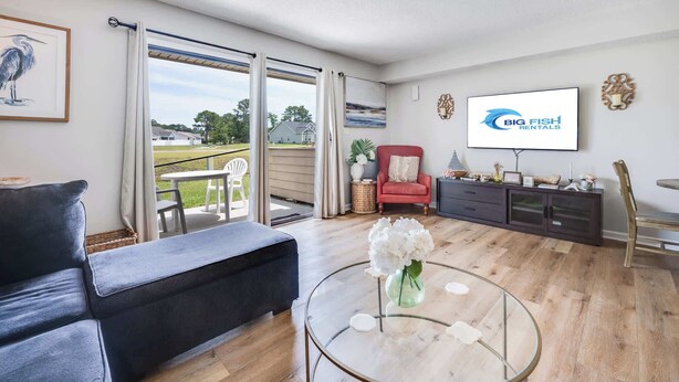 A living room with a sofa, a glass coffee table with flowers, a TV, and a sliding glass door leading to an outdoor area. - 2BR at Grand Palms Resort - Close to Beach (Myrtle Beach)