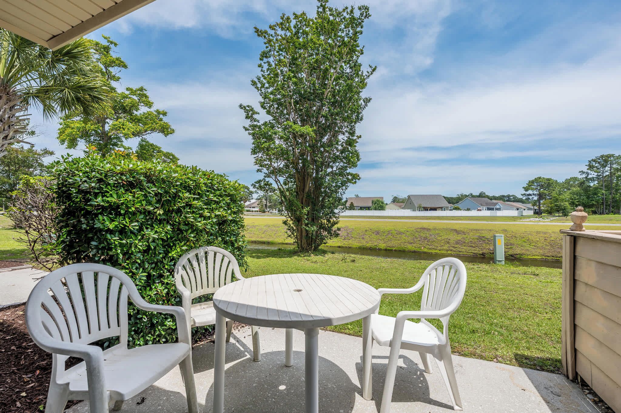A patio with a round table and four chairs, a large tree, and a view of a grassy field and houses.