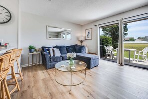 Een moderne woonkamer met een bank, een rond salontafeltje, rieten stoelen, een eettafel en een groot raam met uitzicht op het landschap.