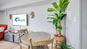 A modern living room with a dining table, chairs, a red sofa, a TV displaying 'BIG FISH RENTALS,' and a large potted plant. - 2BR at Grand Palms Resort - Close to Beach (Myrtle Beach)