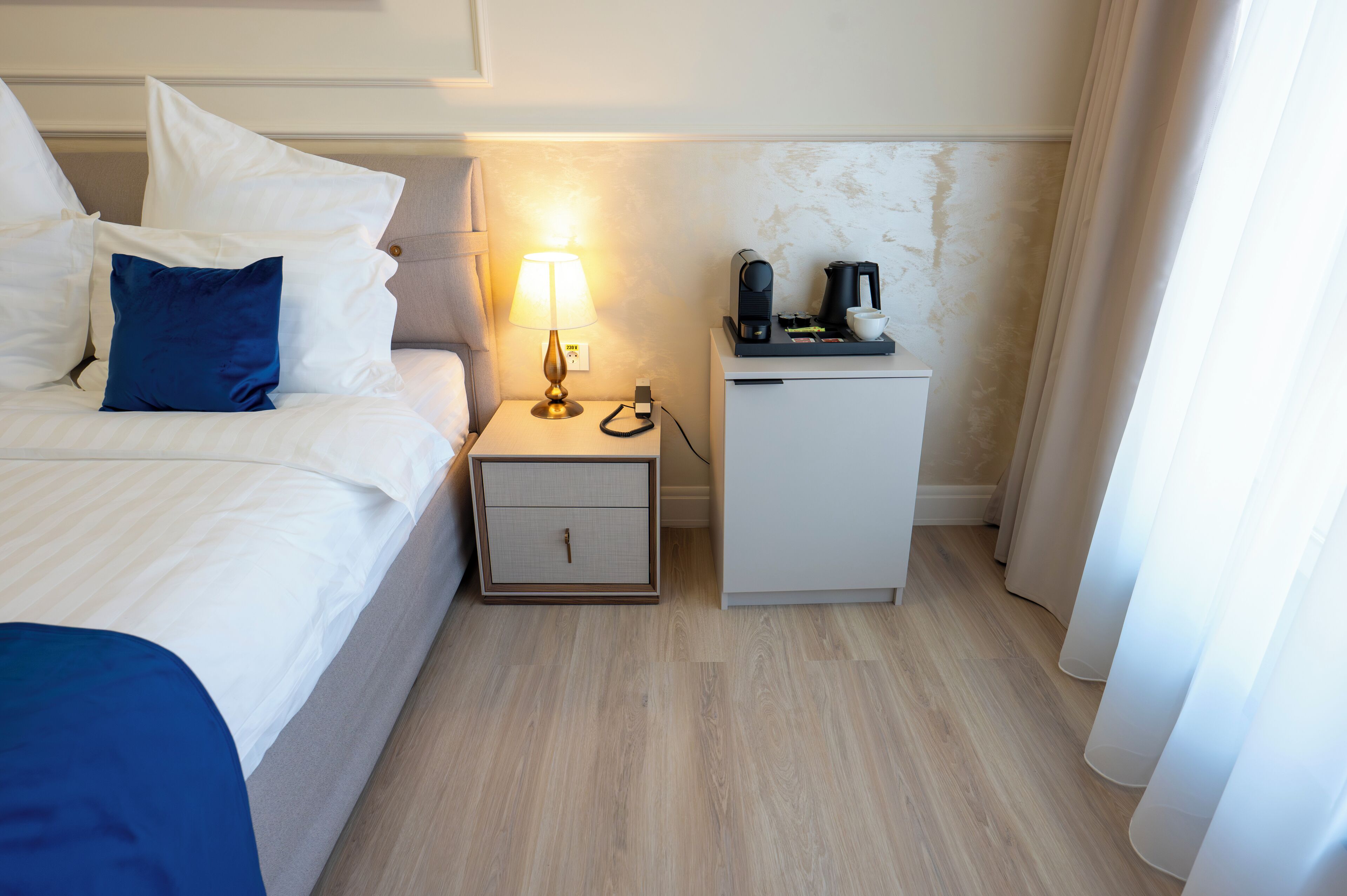 Una habitación de hotel con cama, mesita de noche con lámpara, cafetera y un armario pequeño.