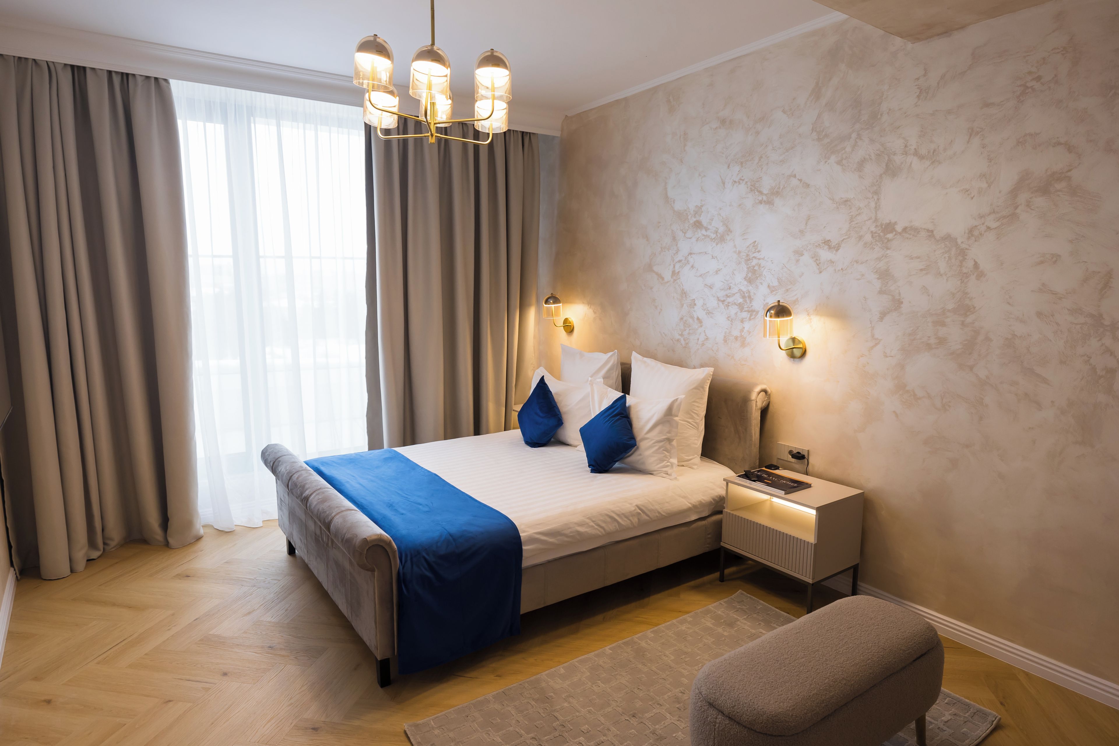 A modern hotel room with a large bed, a bedside table, a reading lamp, and a view of the outdoors.