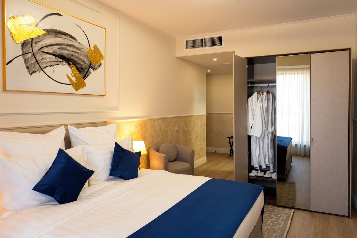 A hotel room with a large bed, a chair, a wardrobe, and a painting on the wall.