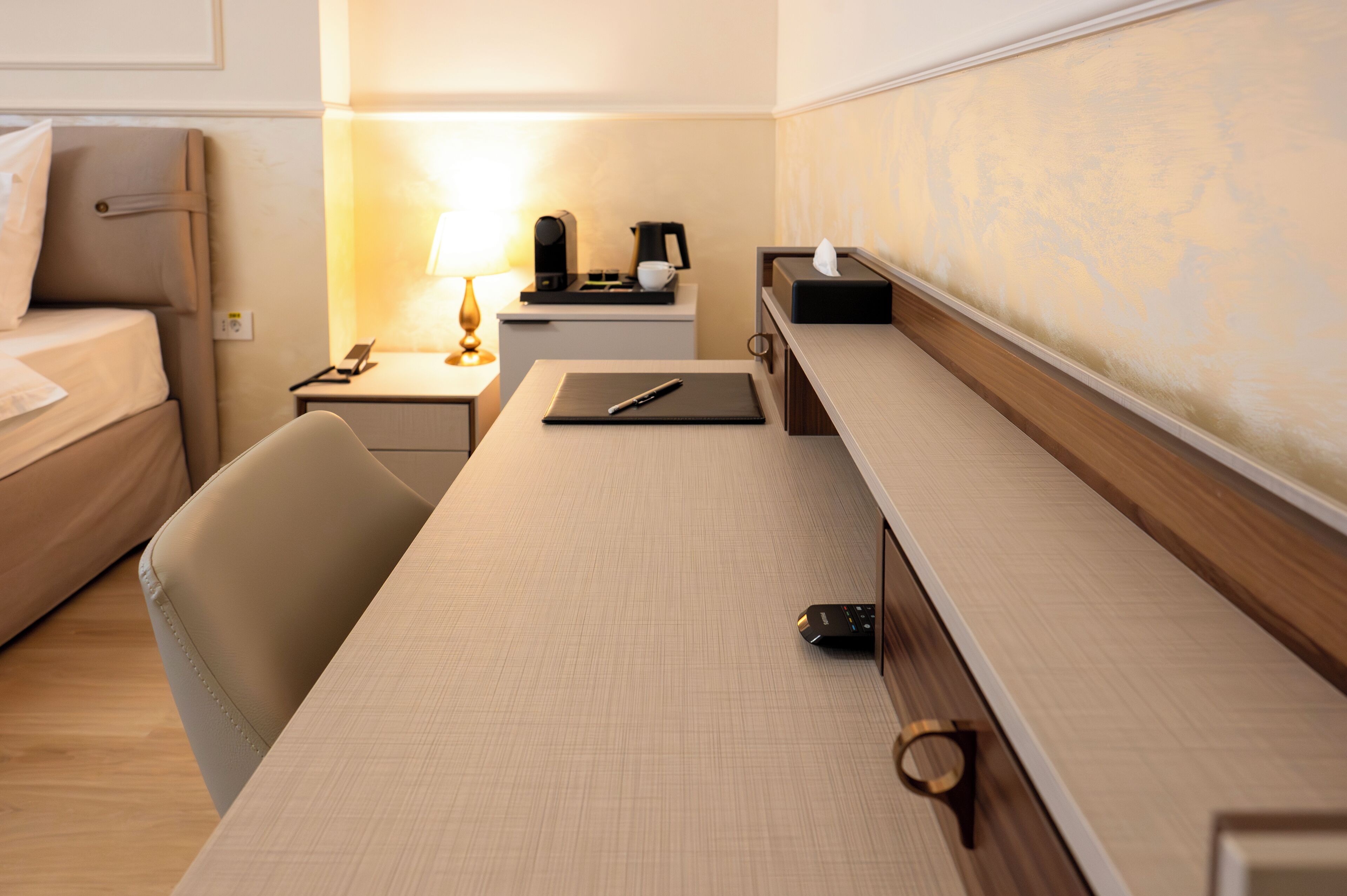 Una habitación de hotel con una cama, un escritorio con lámpara, una cafetera y un control remoto.