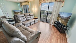 A living room with a beige leather sofa, a flat-screen TV, and a balcony with a scenic view. - 2BR Holiday Towers, Newly Remodeled, Balcony, Pool (Myrtle Beach)