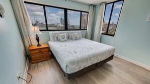 A bedroom with a bed, a nightstand, a lamp, and a view of the city through large windows. - 2BR Holiday Towers, Newly Remodeled, Balcony, Pool (Myrtle Beach)
