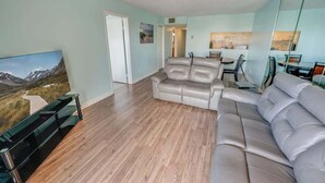 A modern living room with a leather sofa, a flat-screen TV, and a wooden floor. - 2BR Holiday Towers, Newly Remodeled, Balcony, Pool (Myrtle Beach)