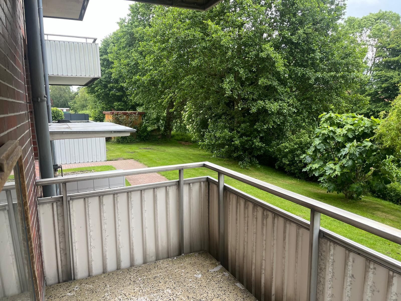 A balcony with a metal railing, a concrete floor, and a view of a green lawn and trees.