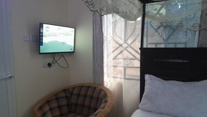 A room with a bed, a television mounted on the wall, a wicker chair, and a window with patterned curtains. - Stan Lodge Babati (Babati)
