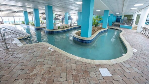 An indoor swimming pool with blue-tiled edges, surrounded by a brick floor and supported by blue pillars. - New Landmark Resort Direct Oceanfront - Low Floor! (Myrtle Beach)