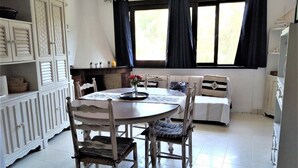 Un coin repas avec une table ronde, des chaises et un banc. On y trouve une fenêtre avec des rideaux, une cheminée et un meuble avec des étagères.