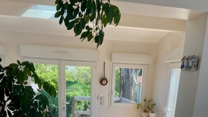 Een ruime kamer met houten vloer, een grote potplant, twee houten stoelen, een witte stoel, en een schuifdeur die leidt naar een buitenruimte met een houten terras.