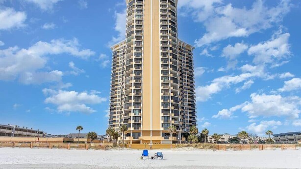A tall, cylindrical hotel with multiple balconies, located near a beach with a sandy shore and palm trees. - Newly Added Maisons, Large Balconies - City View! (Myrtle Beach)