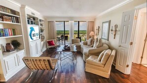 A spacious living room with a large window, a sofa, a coffee table, and a bookshelf. - Newly Added Maisons, Large Balconies - City View! (Myrtle Beach)