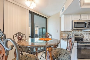 Uma área de refeições com mesa de vidro, cadeiras ornamentadas, cozinha com eletrodomésticos modernos e porta de vidro deslizante que dá para uma varanda.