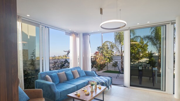 A modern living room with a blue sofa, a wooden coffee table, and a large sliding glass door leading to an outdoor area with plants.