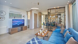 A modern living room with a flat-screen TV, a blue sofa, a wooden coffee table, and a dining area with a chandelier.