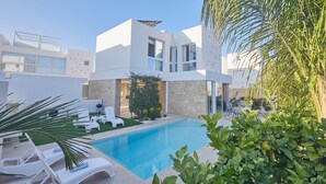 A modern house with a swimming pool, white lounge chairs, and a stone wall.