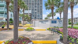 Een aangelegde omgeving met een bordje voor Ocean Forest Plaza, met palmbomen, bloembedden en een verharde looppad.