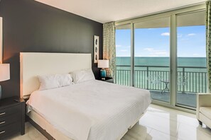 A hotel room with a large bed, a nightstand, a lamp, a view of the ocean through a balcony.